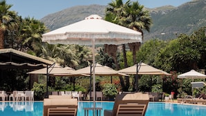 A pool area with lounge chairs and umbrellas, surrounded by palm trees and mountains in the background. - Nia Boutique Hotel by Rapos Resort (Himarë)
