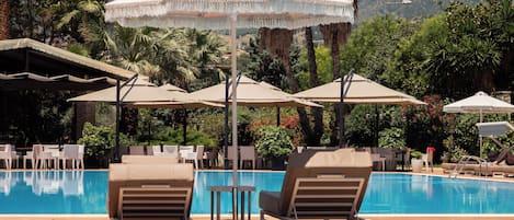 Zona de piscina con sillones y sombrillas, rodeada de palmeras y montañas al fondo.