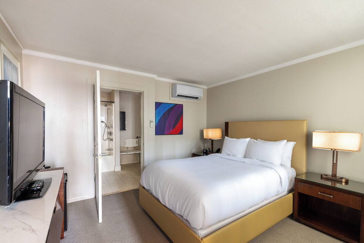 A hotel room with a bed, bedside tables, a TV, and an air conditioning unit.
