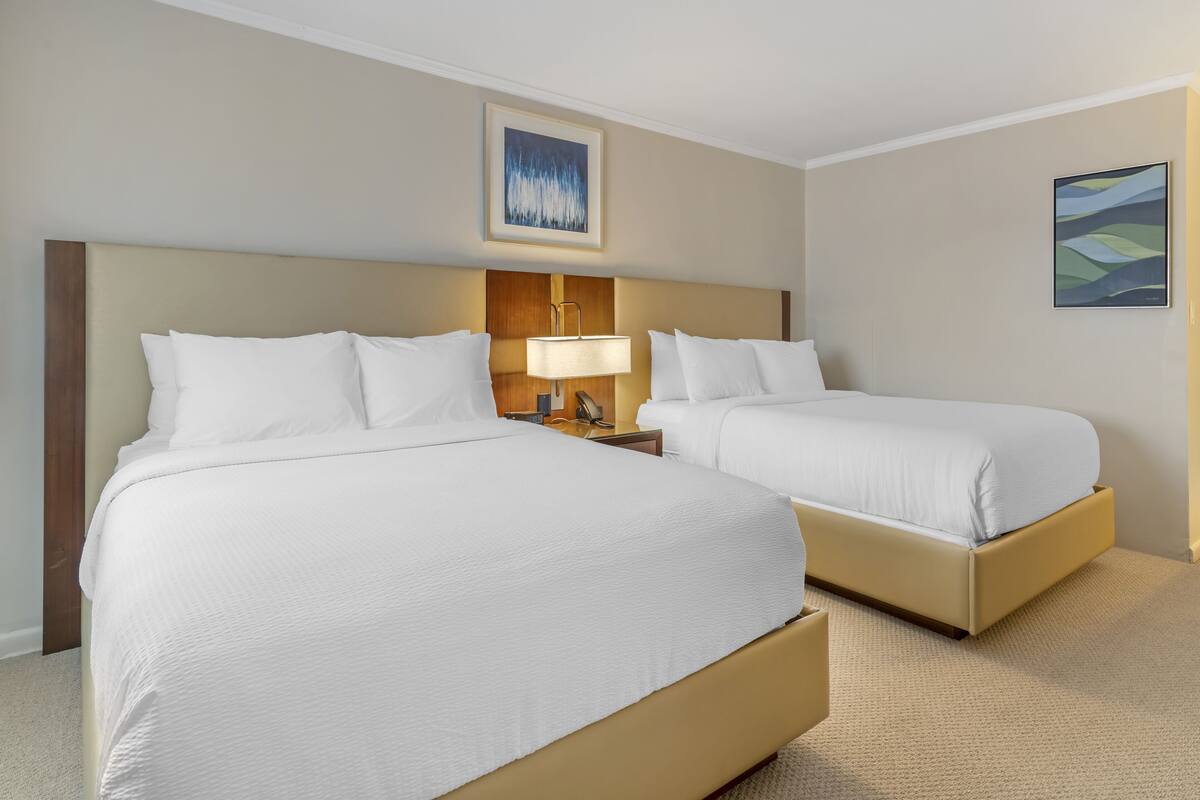 A hotel room with two beds, a nightstand with a lamp, and artwork on the walls.