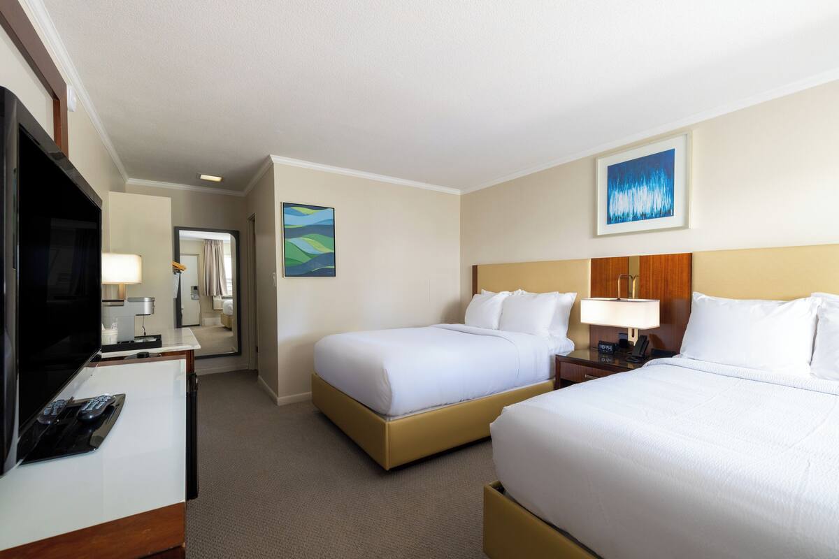A hotel room with two beds, a desk with a TV and remote, a lamp, and a painting on the wall.