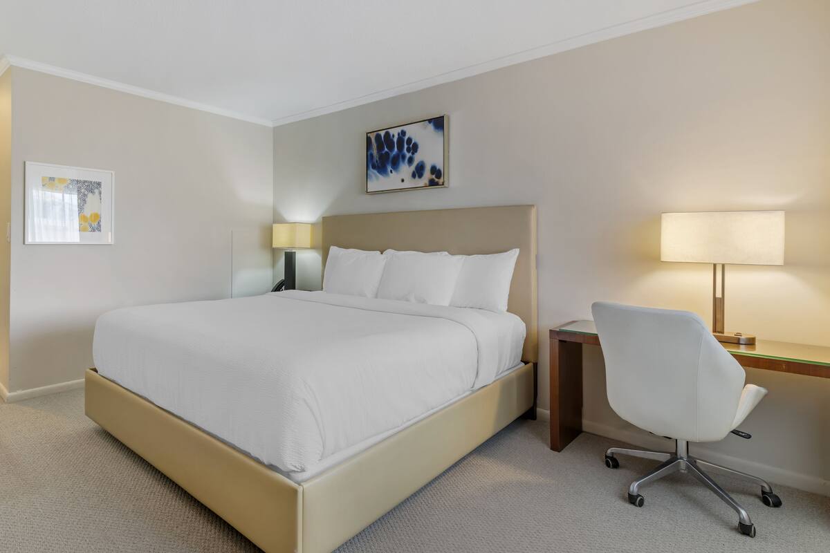A hotel room with a bed, a desk, a chair, a lamp, and two wall art pieces.
