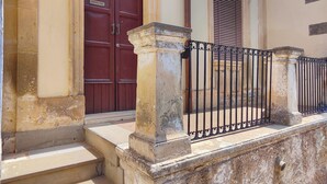 Un bâtiment avec une porte rouge et une fenêtre à volets, un escalier en pierre et une rambarde en métal.