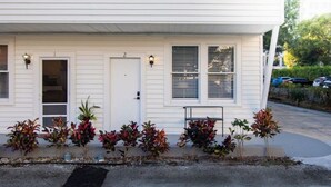 A white two-story house with a front door, two windows with shutters, and a small bench. - The Downtown Hibiscus House (West Palm Beach)