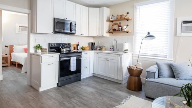 A modern kitchen with white cabinets, a black stove, and a microwave. There is a dining area with a table and chairs, and a window with blinds. - The Downtown Hibiscus House (West Palm Beach)