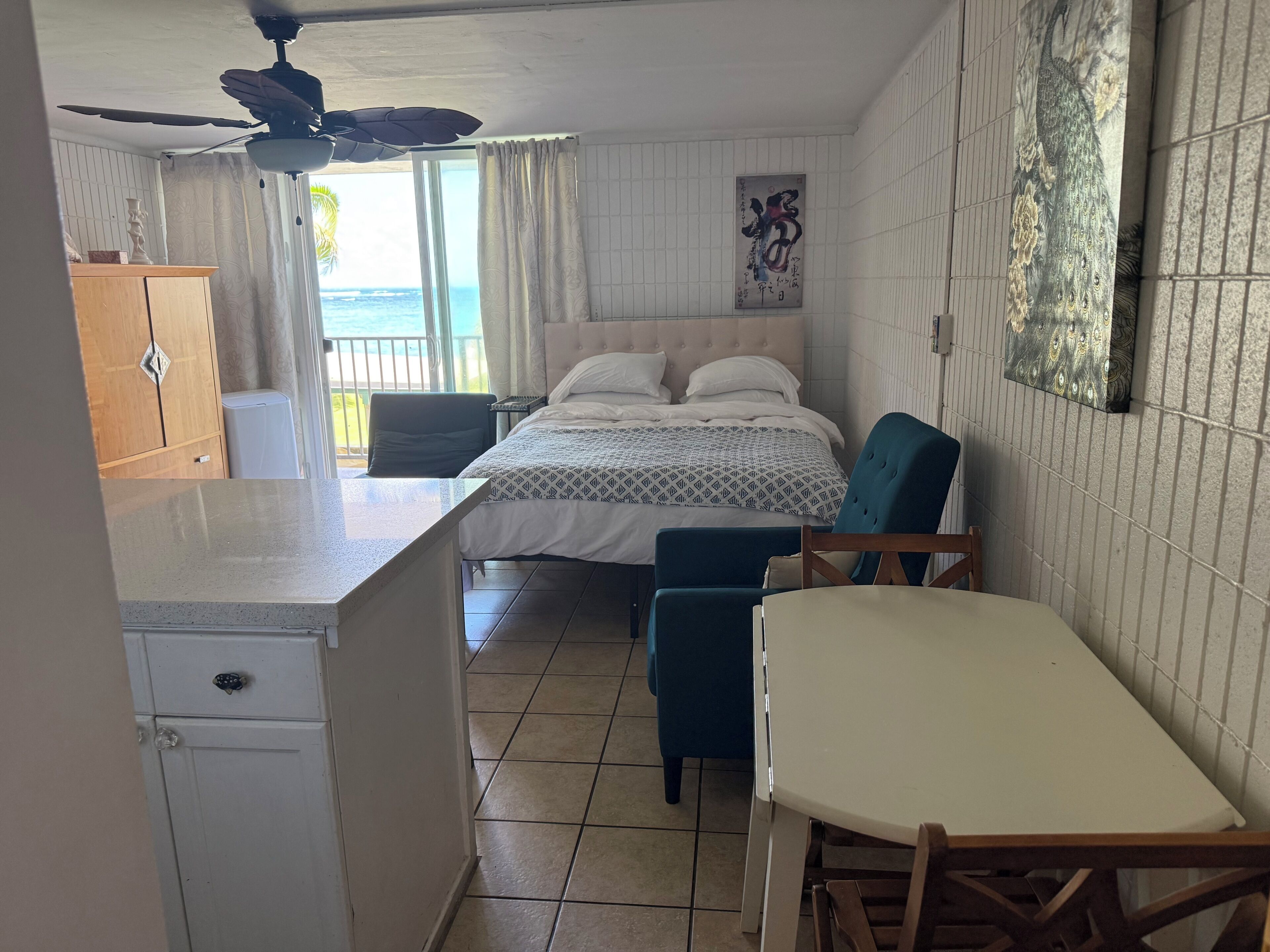 Un dormitorio con cama, ventilador de techo, ventana con cortinas, una obra de arte en la pared y vista al océano.