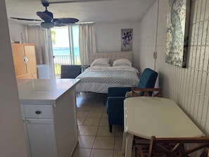 A bedroom with a bed, a ceiling fan, a window with curtains, a wall-mounted artwork, and a view of the ocean. - Turtle View Beachfront, snorkeling, free parking, wash-dryer in unit, AC. (Hauula)