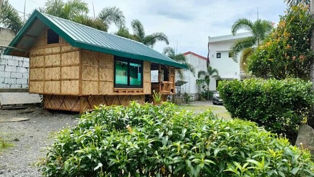 A small wooden house with a green roof, surrounded by greenery and a white building in the background.