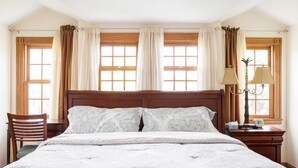 Une chambre avec un grand lit, deux fenêtres avec des rideaux, une table de chevet avec une lampe et une chaise.