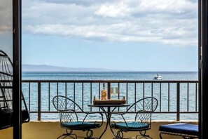 A balcony with two chairs, a small table with a bottle and glasses, and a view of the sea with a boat in the distance.