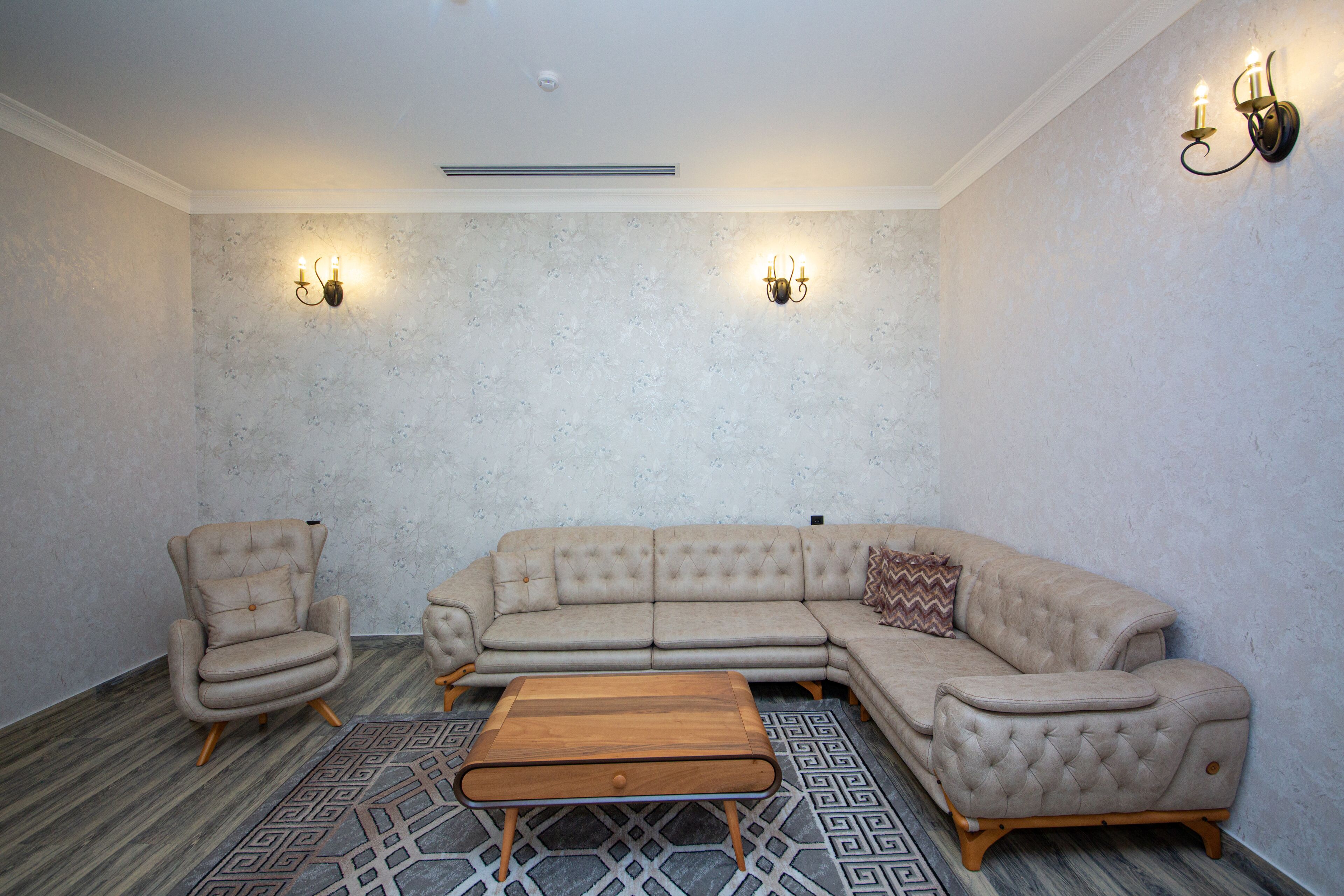 A room with a beige sectional sofa, a single armchair, and a wooden coffee table. There are two wall-mounted sconces providing light.