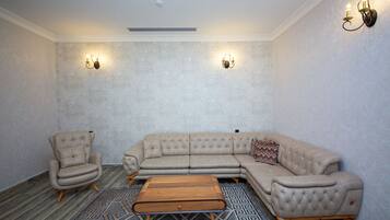 A room with a beige sectional sofa, a single armchair, and a wooden coffee table. There are two wall-mounted sconces providing light.