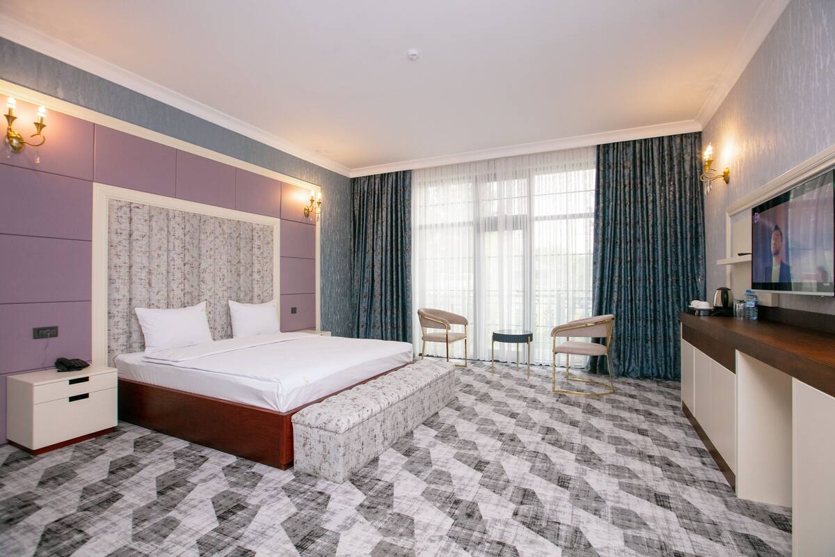 Ein Hotelzimmer mit einem großen Bett, einem Nachttisch, einem kleinen Schreibtisch mit Stuhl, einem Fernseher und einem Fenster mit Vorhängen.