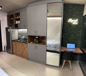 Quarto compacto com cozinha compacta, uma cama e uma escrivaninha com um laptop.