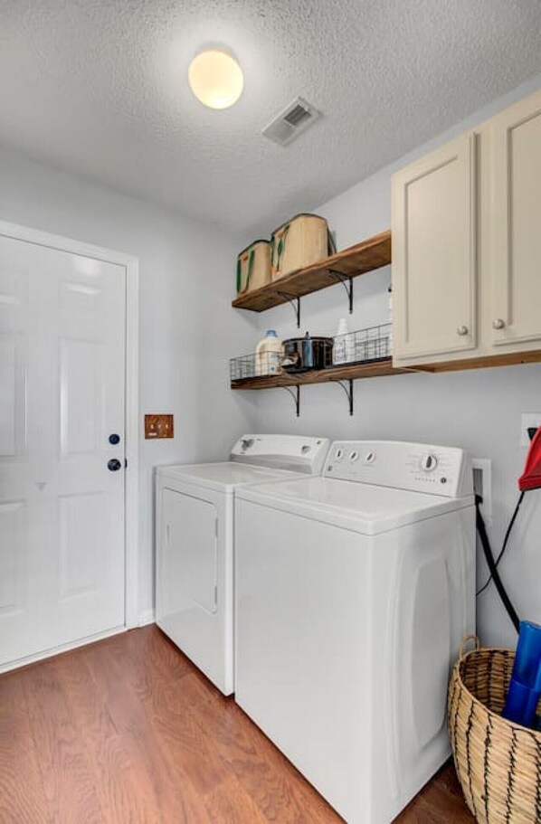 A laundry room with a washing machine, a dryer, wooden shelves above, a door, and a basket. - House with Backyard near the Charleston Naval Base (North Charleston)