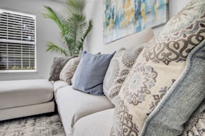 Un salon avec un canapé, une fenêtre avec des stores, une plante et un tableau accroché au mur.