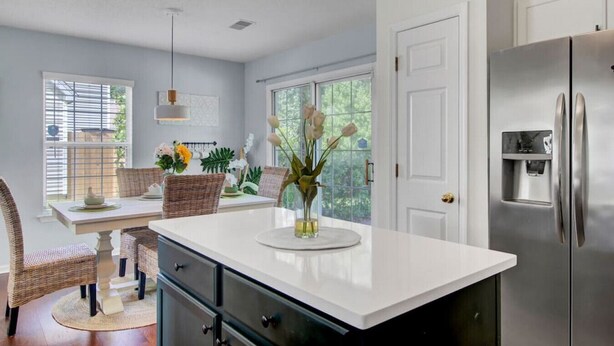 A kitchen with a white countertop, stainless steel refrigerator, wicker chairs, and a dining table with a vase of flowers. - House with Backyard near the Charleston Naval Base (North Charleston)