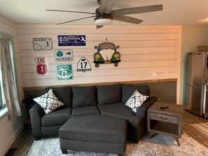 A room with a gray sectional sofa, a small wooden nightstand, and a ceiling fan. The walls feature various road signs and a welcome sign for Michigan.