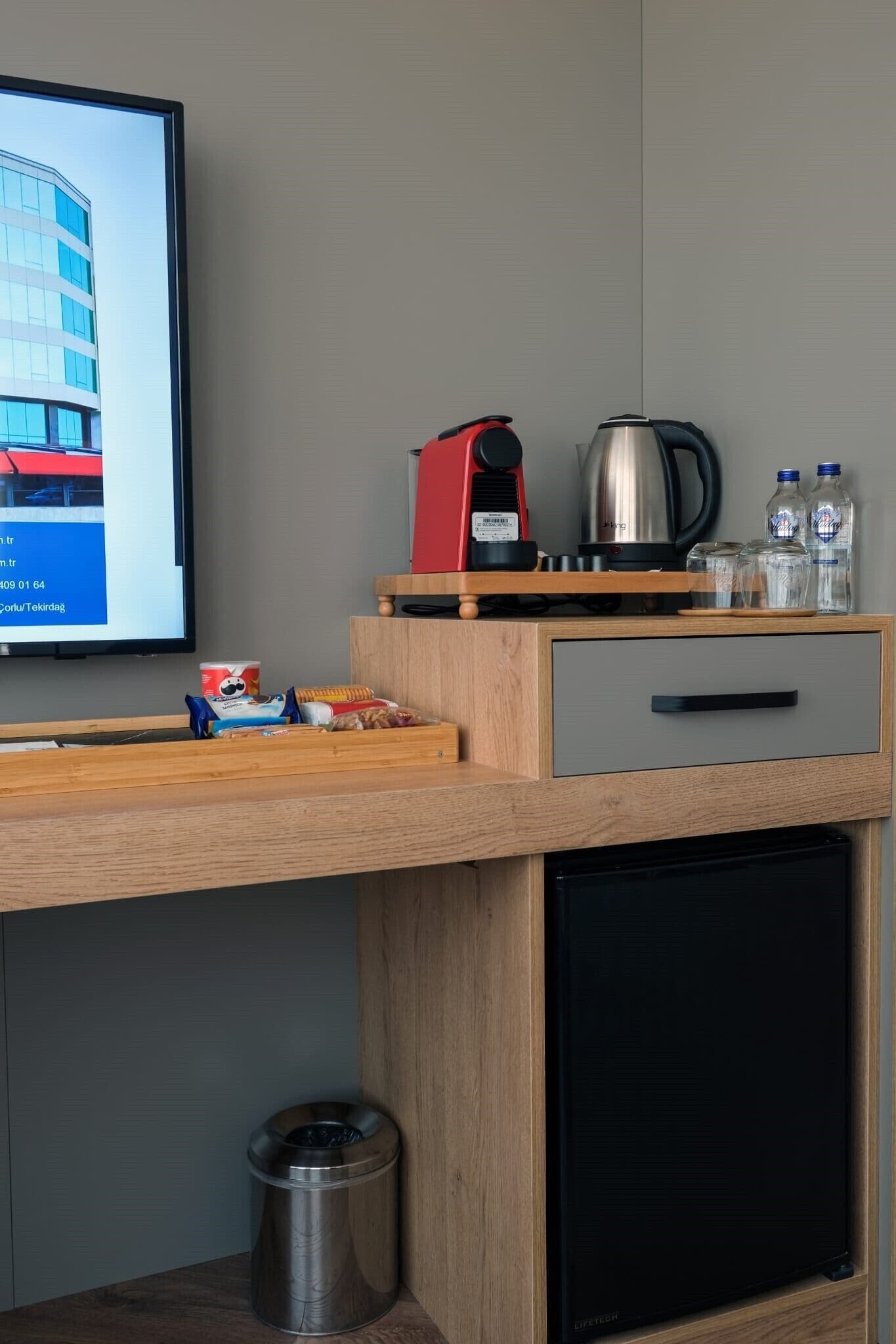 A hotel room with a wooden desk, a coffee maker, a kettle, a trash bin, and a television displaying a building.