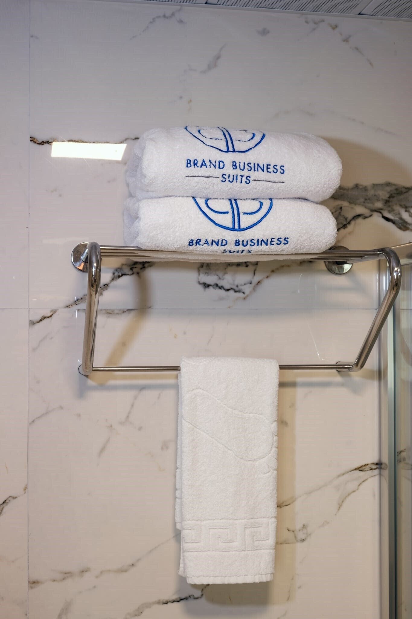 Un baño con pared de mármol, un perchero con dos toallas de marca y una toalla lisa.