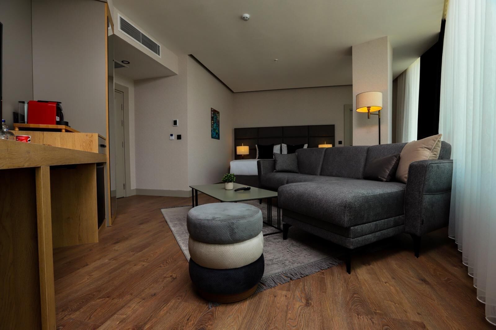 A modern living room with a grey sofa, a wooden coffee table, and a small ottoman. There is a red coffee machine on a wooden counter.
