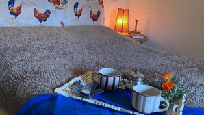 Una habitación con una cama cubierta con una manta azul, una bandeja con una taza, una pequeña vela y un arreglo floral. Hay una pared con un estampado de gallo y una lámpara de pared.