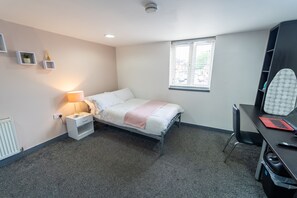 A bedroom with a bed, bedside table with lamp, window, and desk with laptop.