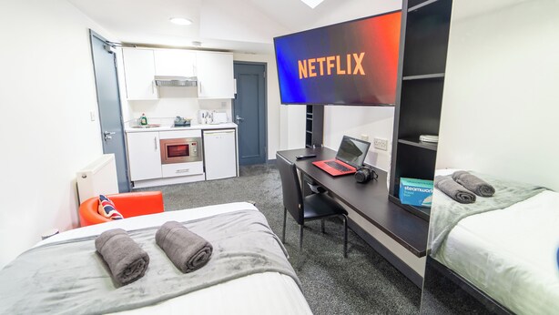 Quarto de hotel com duas camas, uma mesa de trabalho com um laptop e uma câmera, uma TV com o logotipo da Netflix, uma cozinha compacta e vista para o exterior.