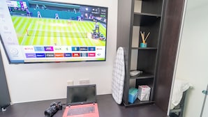 Quarto de hotel com escrivaninha, apresentando um laptop, uma câmera e uma TV de tela plana exibindo uma partida de tênis.