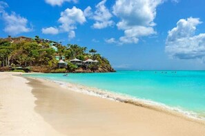 A beach with clear turquoise water, a sandy shore, and a building with multiple structures on a hill.
