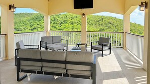A covered outdoor seating area with a wooden ceiling, a flat-screen TV, and a view of a forested hillside.