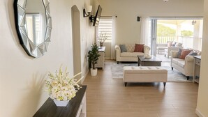 A spacious living room with a sofa, ottoman, and a coffee table. There is a potted plant on a console table and a large mirror on the wall.