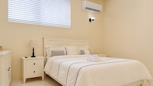 Un dormitorio con cama, mesitas de noche, lámpara, aire acondicionado instalado en la pared y una ventana con persianas.
