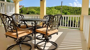 Un patio techado con una mesa y sillas, barandal blanco y vista a un paisaje verde.