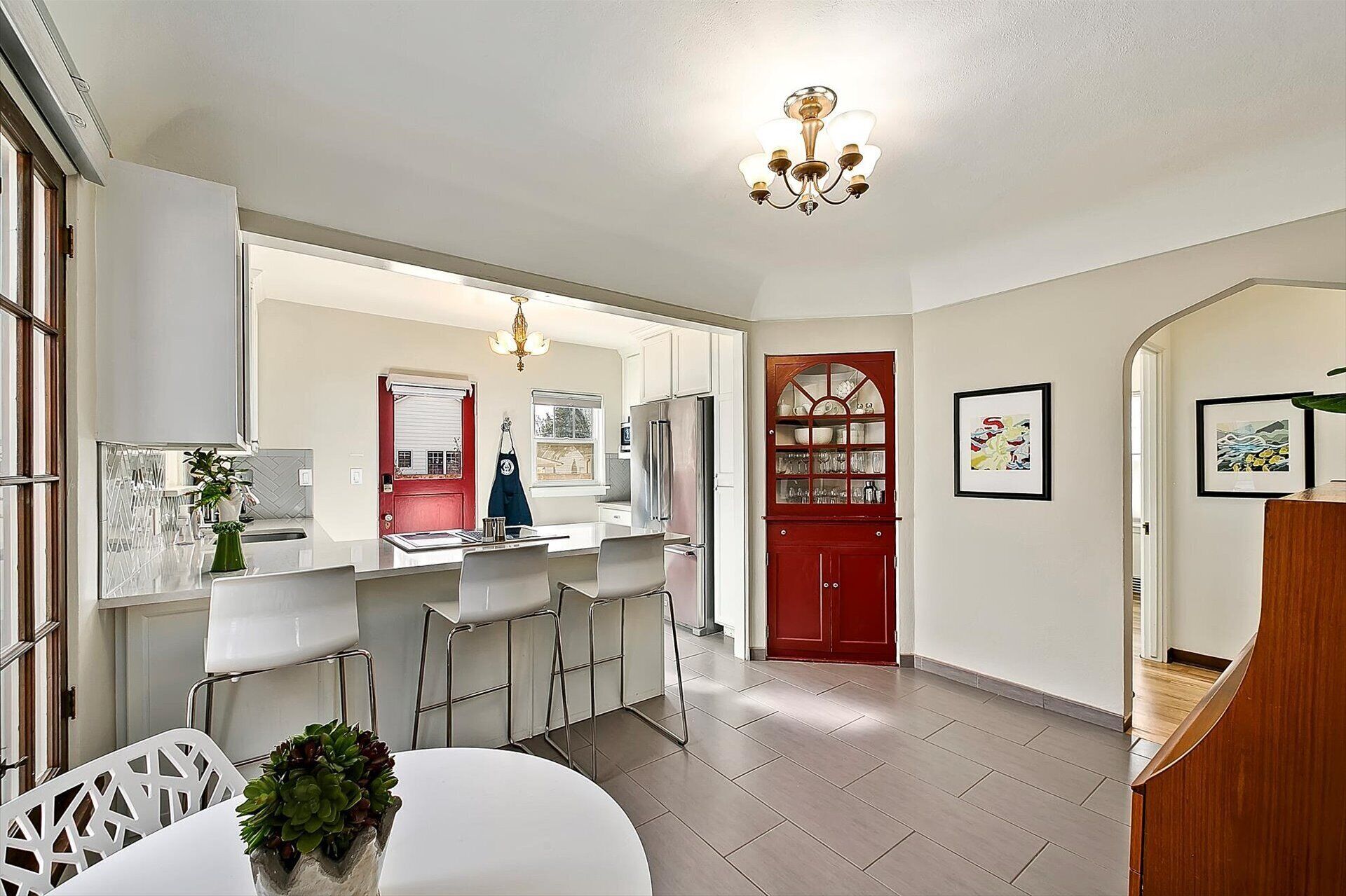 Une cuisine moderne avec des meubles blancs, une porte rouge et un coin repas comprenant une table et des chaises blanches.