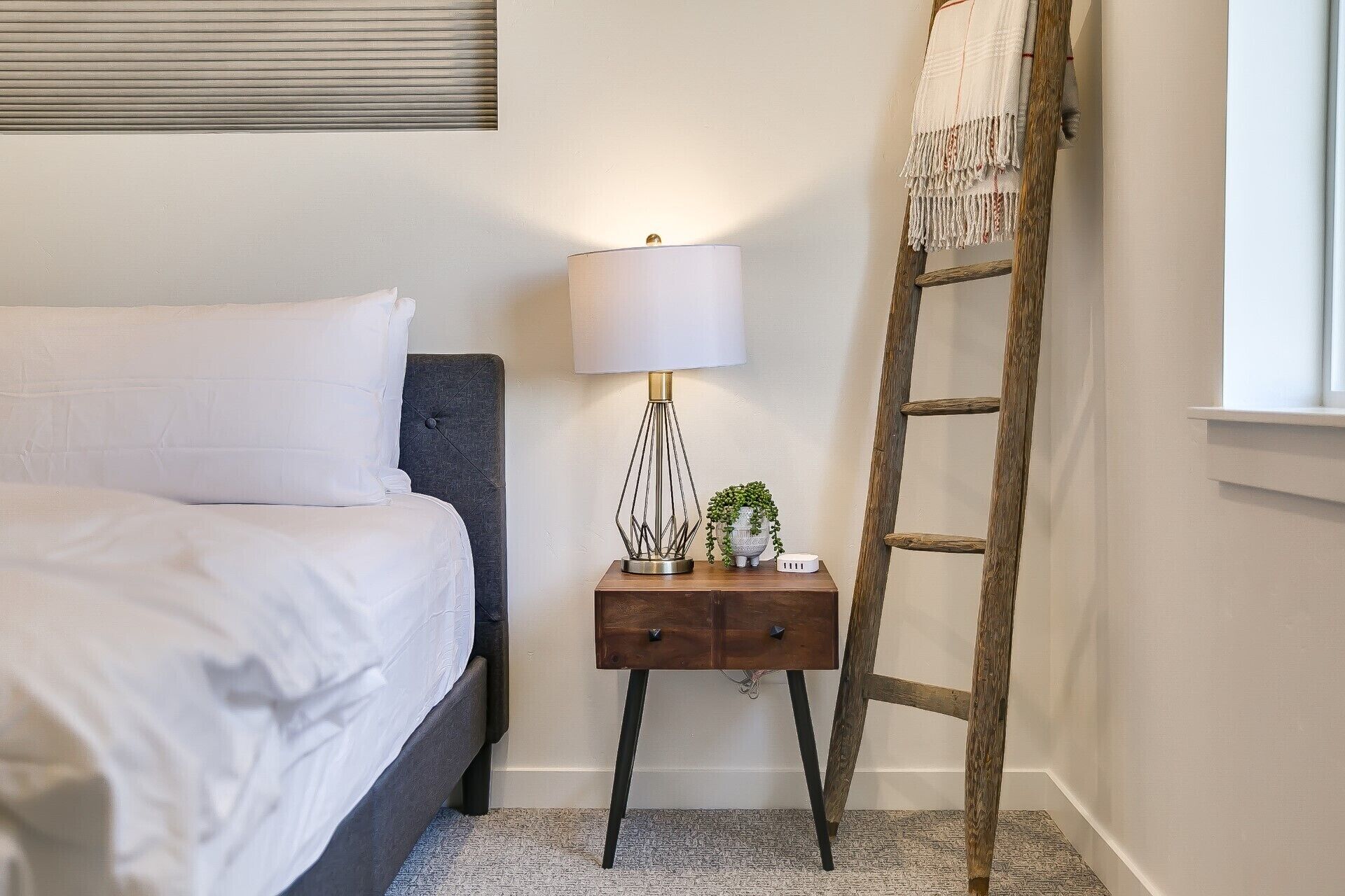 A neatly made bed with white linens, a bedside lamp, a small wooden nightstand with a plant, and a wooden ladder leaning against the wall.