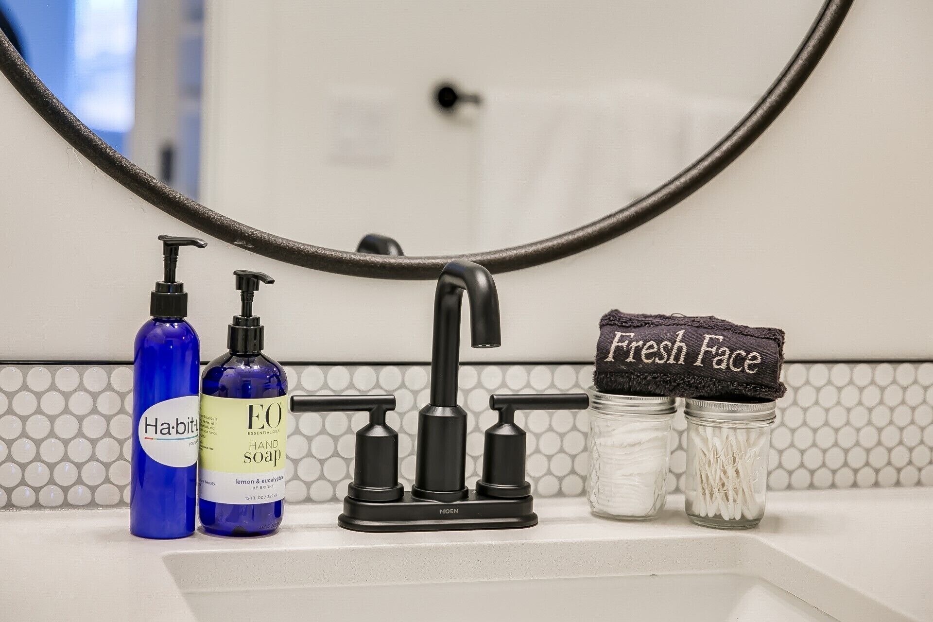 Un lavamanos con grifo negro, dos botellas de jabón para manos y un frasco de hisopos con la etiqueta 'Fresh Face'.