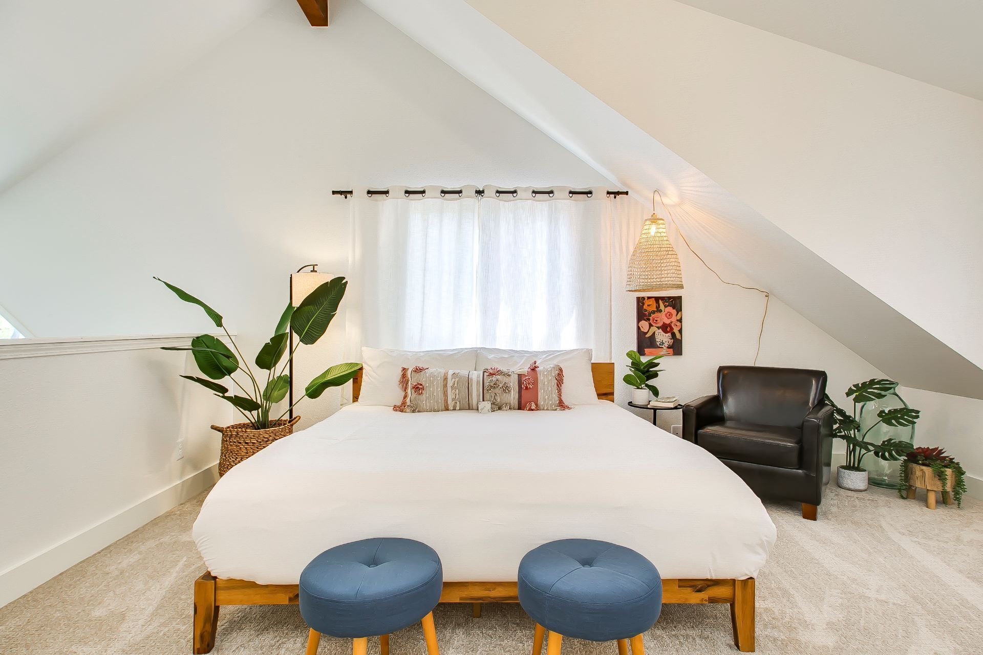 A bedroom with a large bed, two blue ottomans, a black leather chair, and several potted plants.