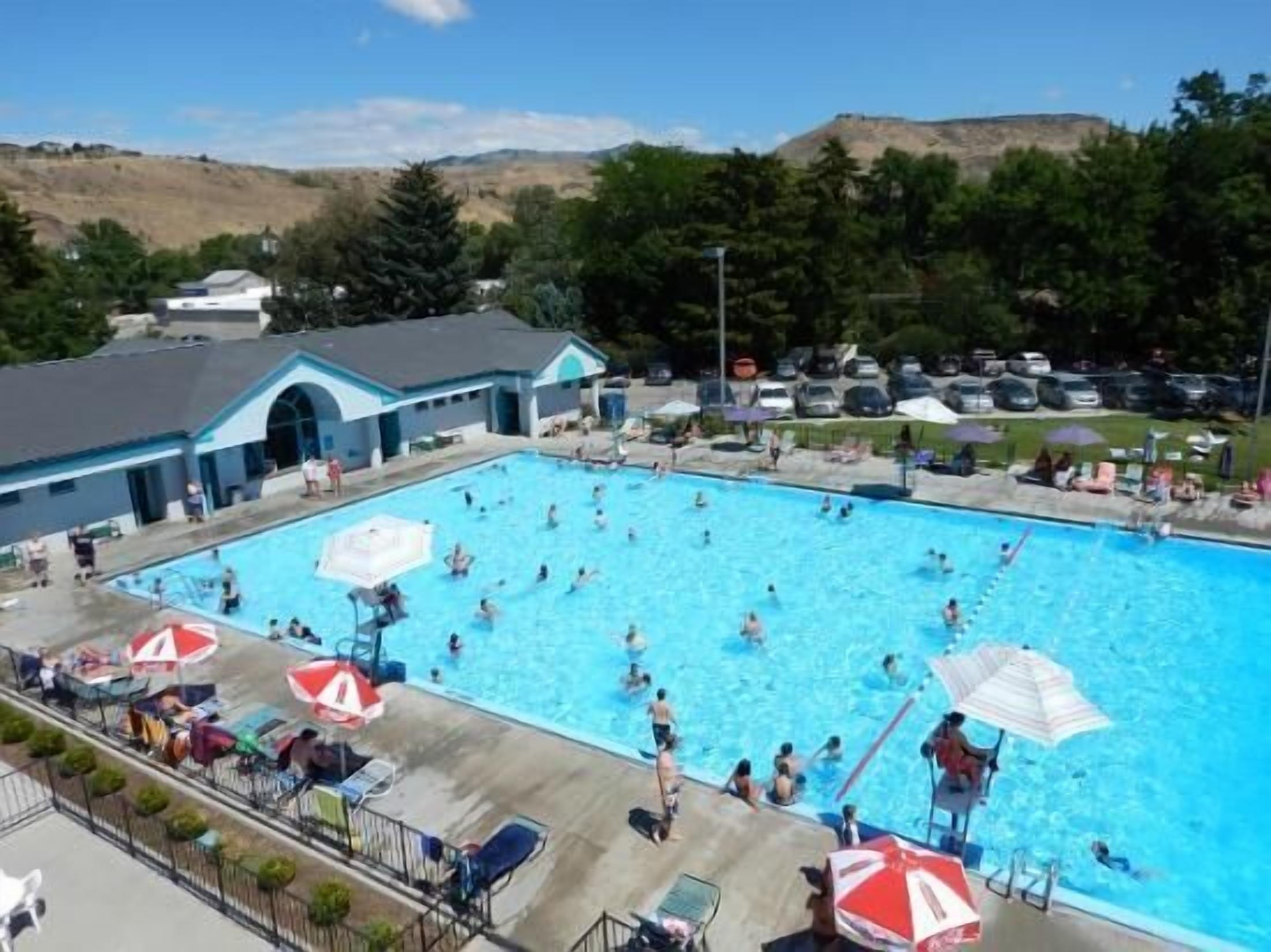 Una piscina con gente, sombrillas y un edificio de techo azul.
