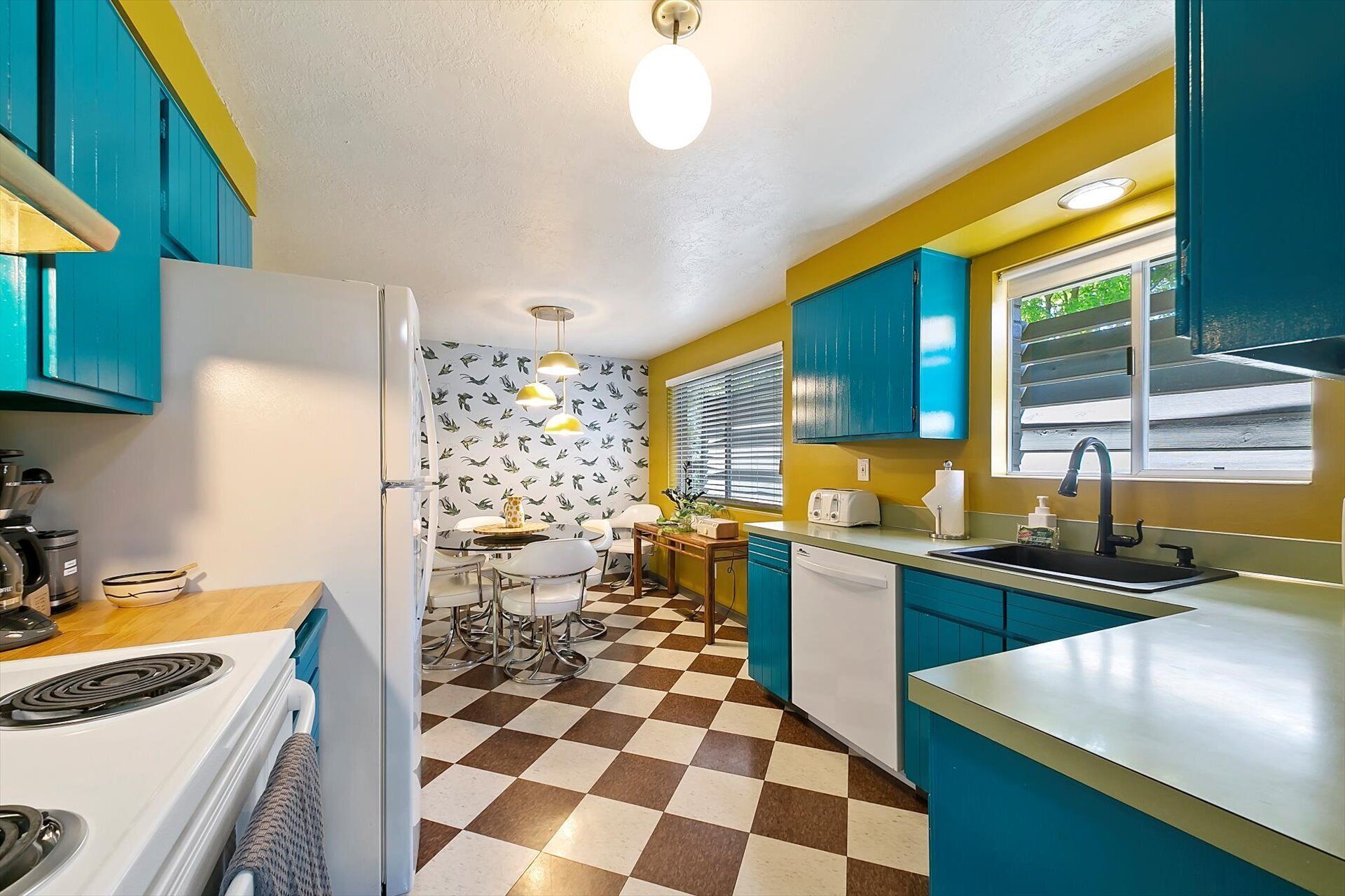 Een keuken met blauwgroene kastjes, een geruit vloertje en een raam met jaloezieën.