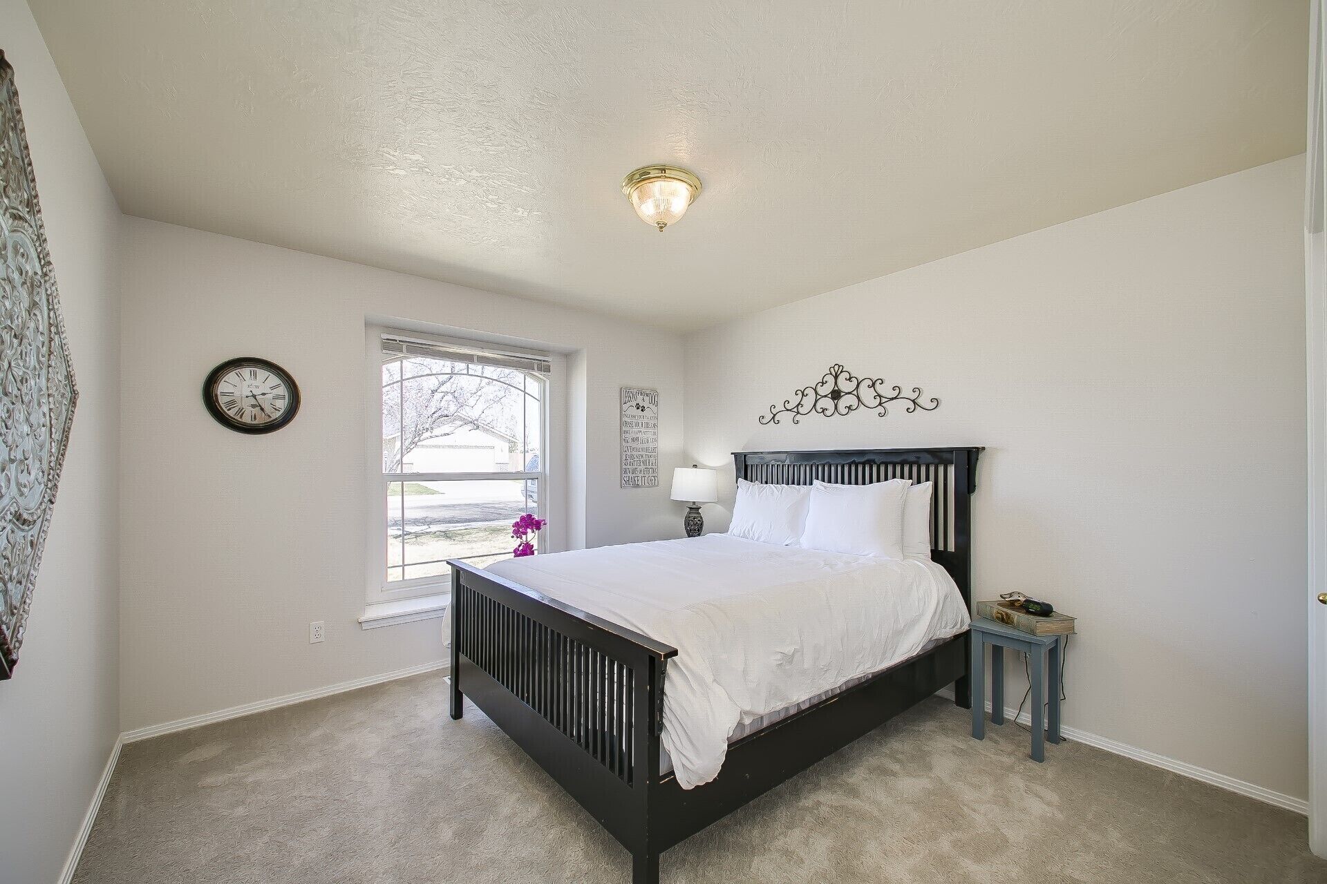 Une chambre à coucher comprenant un lit, une table de chevet, une horloge murale, une fenêtre et un luminaire au plafond.