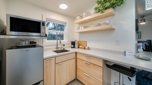 A modern kitchen with a built-in microwave, stainless steel refrigerator, and wooden cabinets. There is a window providing natural light and a plant on a shelf above the counter. - The Green Spot Studio (Boise)