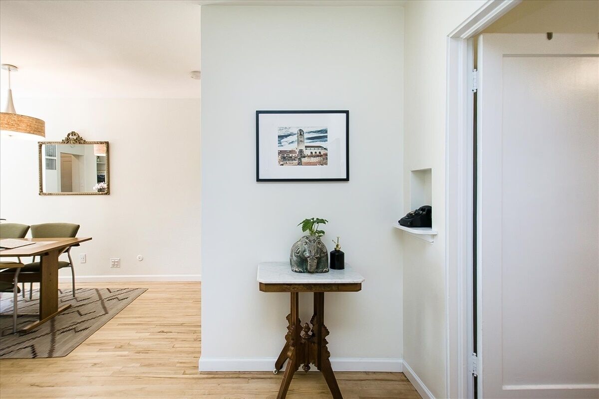 A room with a dining table, a mirror, a framed picture on the wall, and a small table with a plant and a vase.