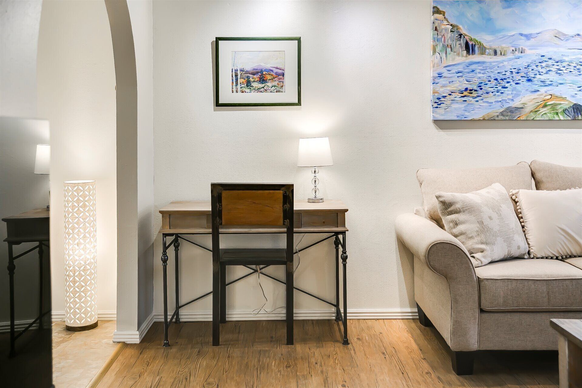 A room with a wooden desk, a cushioned chair, a sofa, and a painting on the wall.