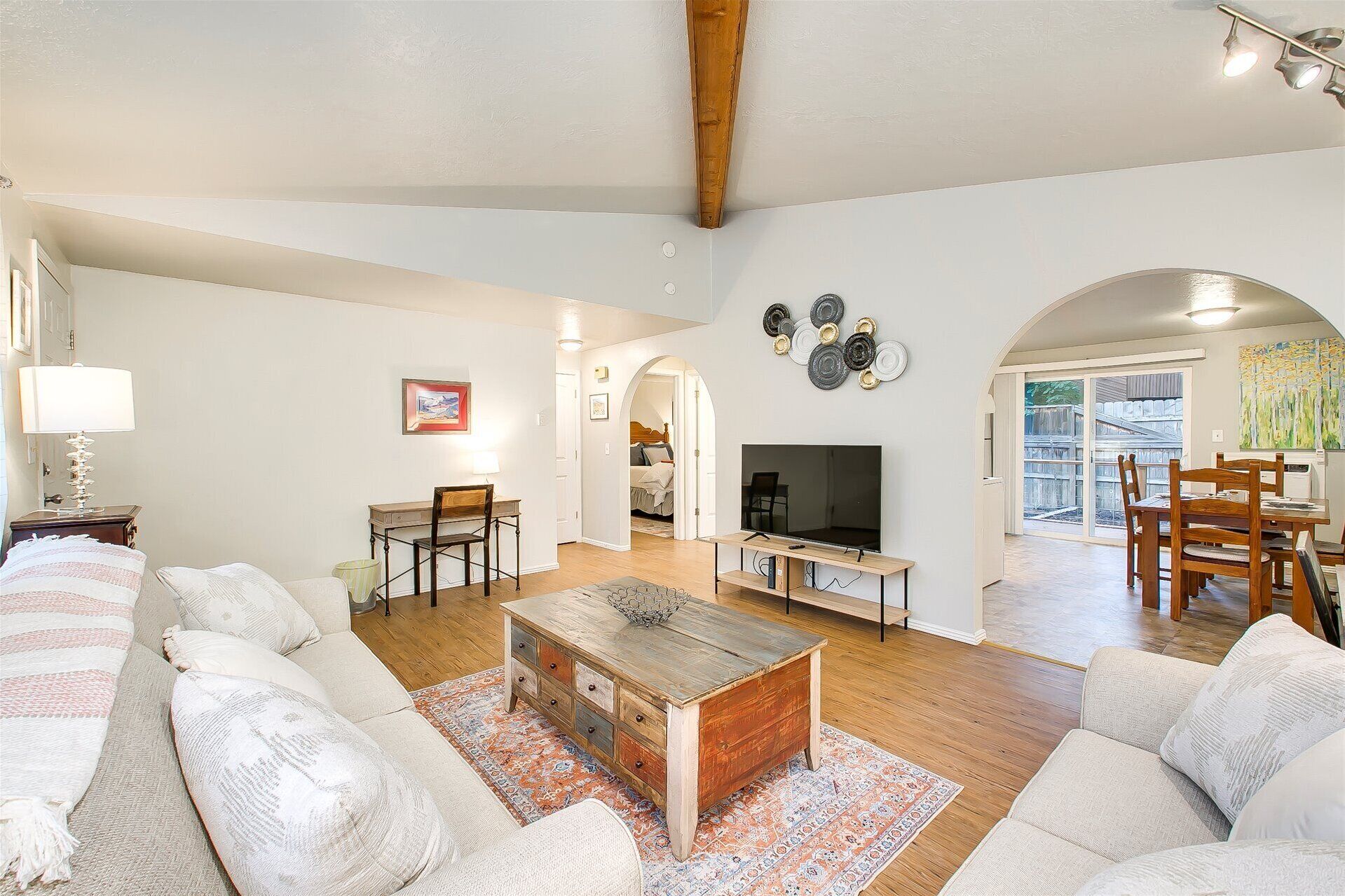 A living room with a sofa, coffee table, TV, and dining area. There is a painting on the wall and a window with a view.
