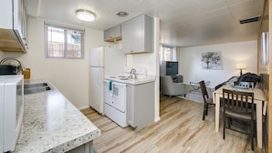 Cocina compacta con armarios blancos, un refrigerador, una estufa y un lavabo. Hay un comedor con una mesa y sillas de madera.