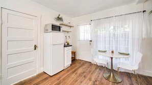 Cocina compacta con muebles blancos, microondas y una pequeña isla con pileta. Hay una mesa de comedor con sillas blancas y una ventana con cortinas traslúcidas.