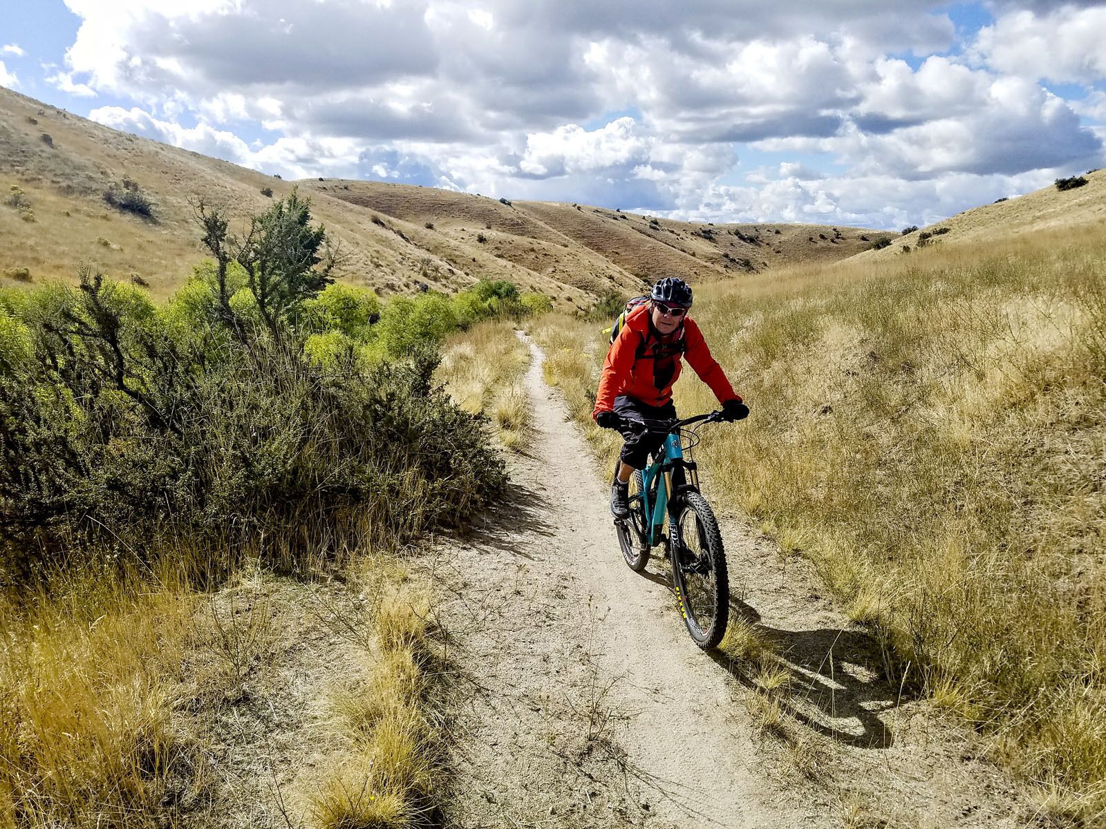 Une personne fait du vélo tout terrain sur un sentier accidenté, entourée d’herbes sèches et d’une végétation clairsemée.