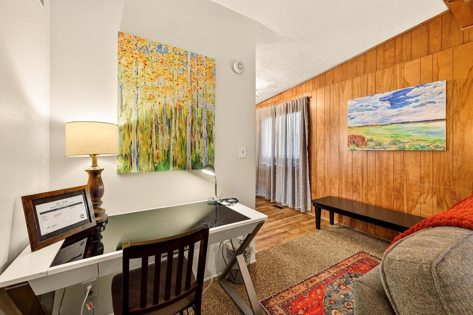 A room with a desk, chair, lamp, paintings, and a couch.