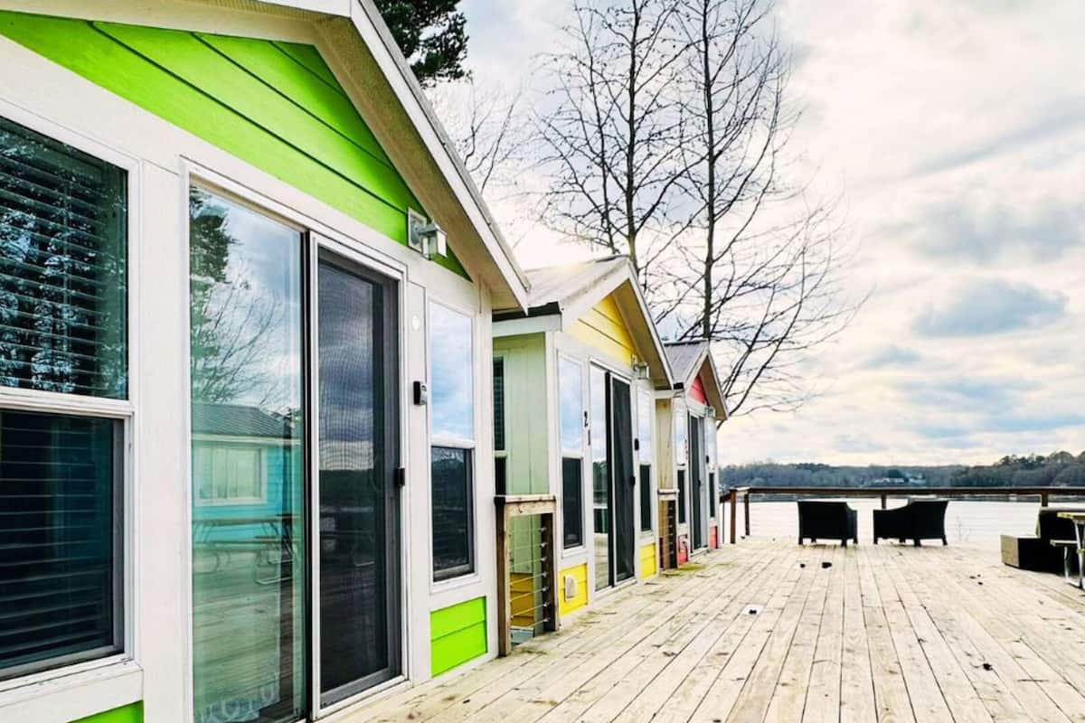 A row of cabins with large windows and sliding doors, a wooden deck, and a view of a lake.